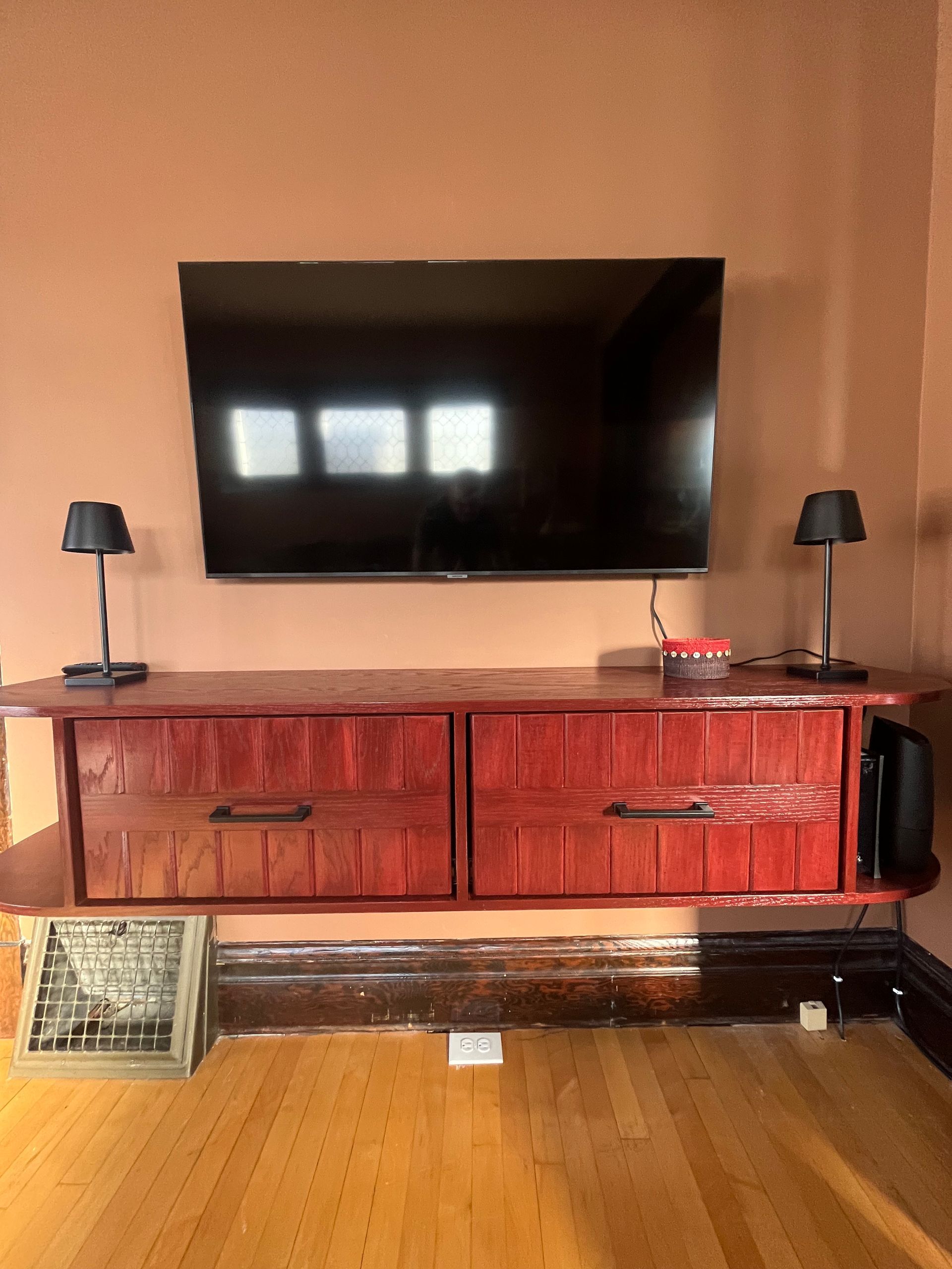 A wall-mounted TV sits above a long, rustic red wood cabinet with two drawers and black lamps on either side.