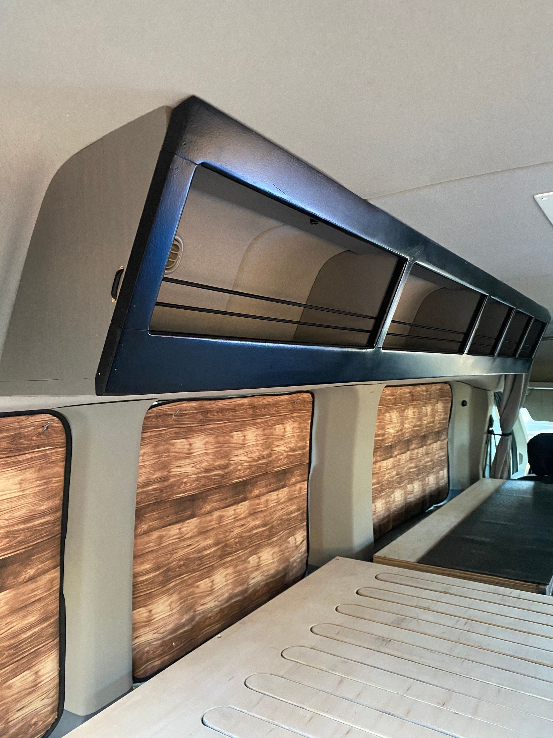 Interior view of a camper van with overhead storage cabinets, faux-wood window covers, and a light-colored platform bed.