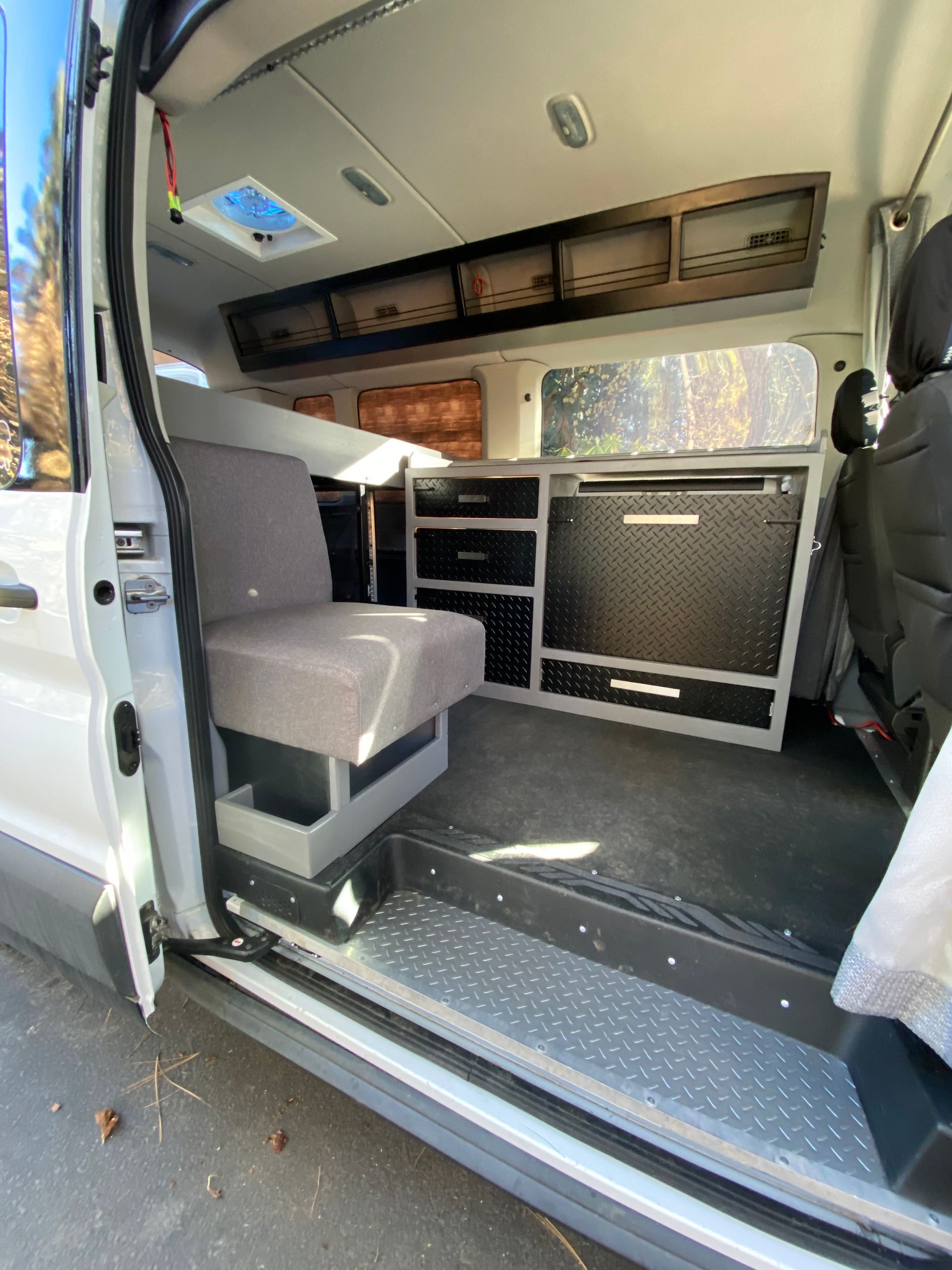 Interior of a van conversion featuring a grey jump seat, metal cabinetry, and a textured floor step near the open door.