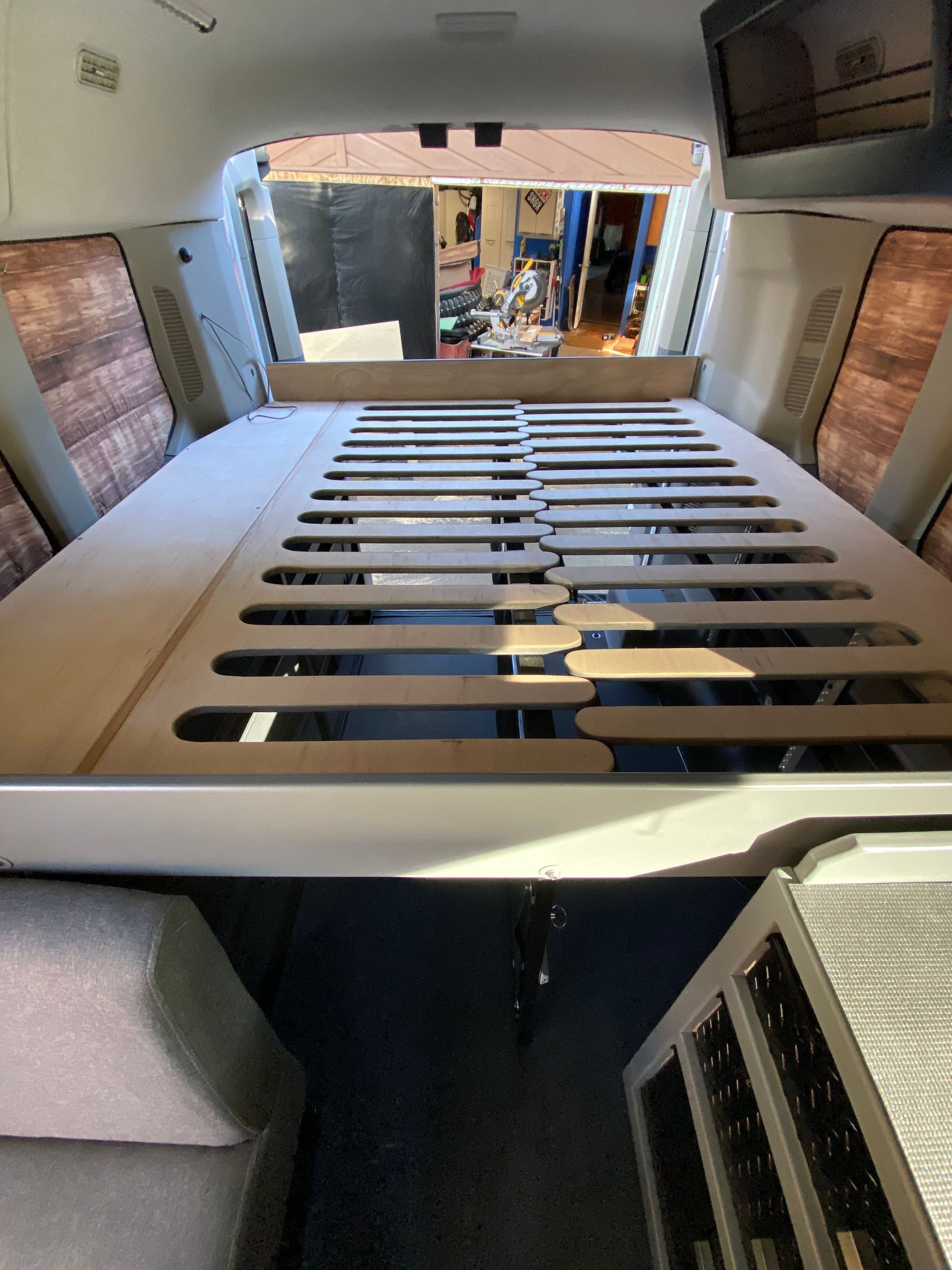 The interior of a camper van showing a raised bed platform with slatted supports, wooden paneling, and beige upholstery.