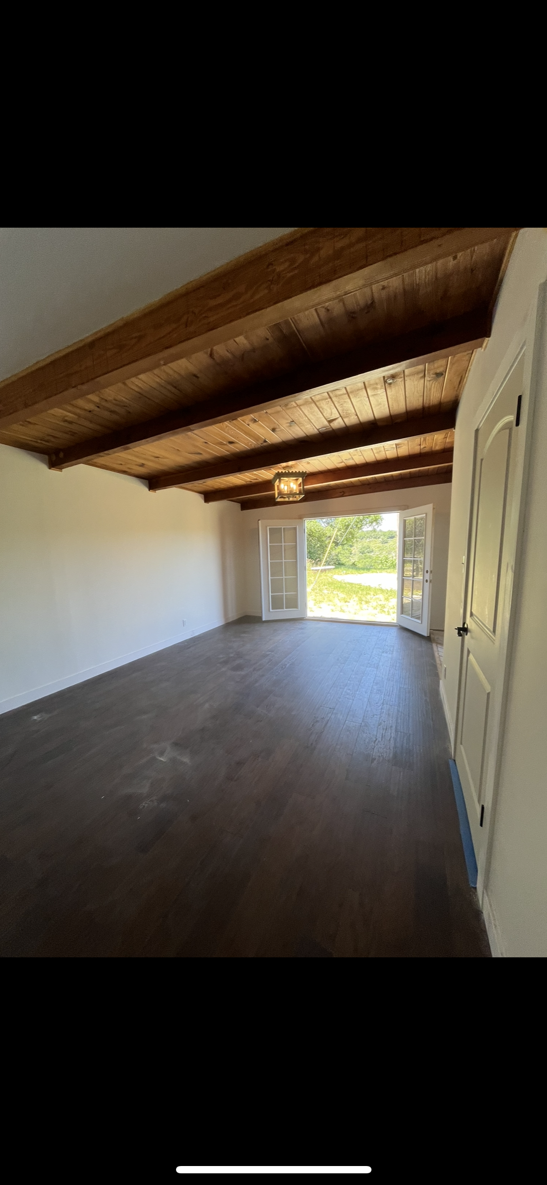 An empty room with dark wood flooring, white walls, exposed wooden ceiling beams, and French doors leading to a yard.
