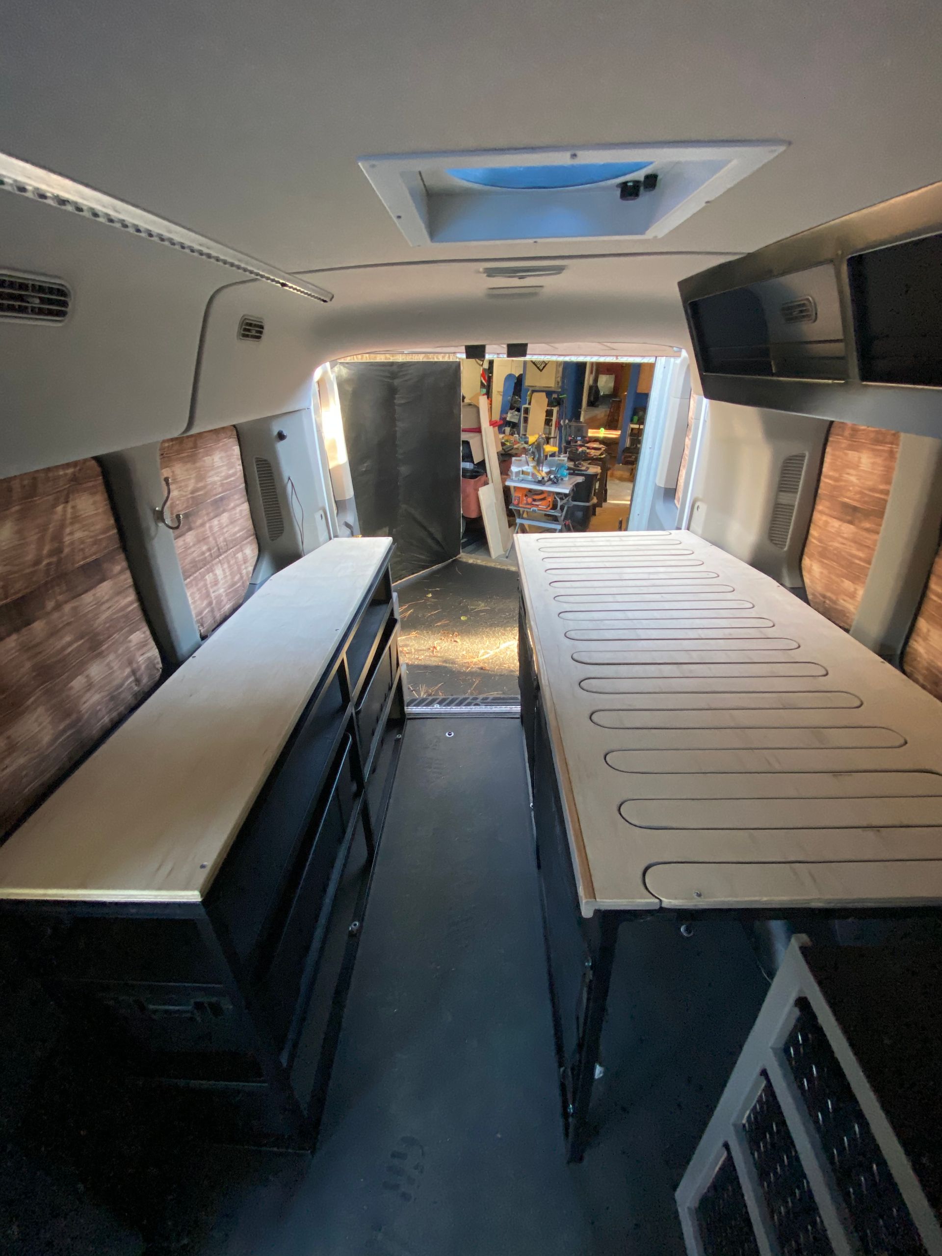 Interior view of a van conversion featuring two long wooden platforms with storage underneath, leading toward open doors.