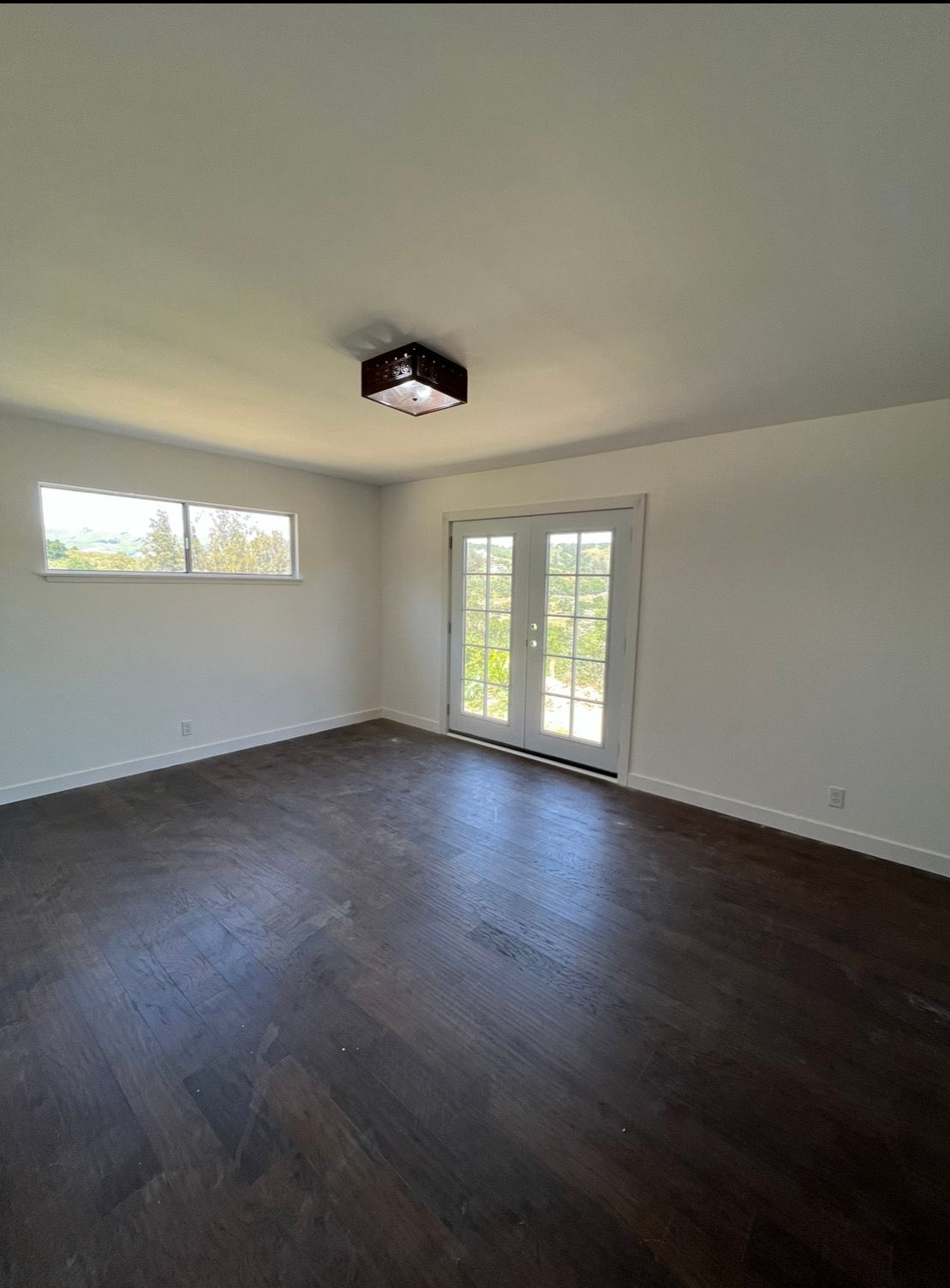 A minimalist, unfurnished room featuring dark wood herringbone flooring, white walls, a square ceiling light, and windows.