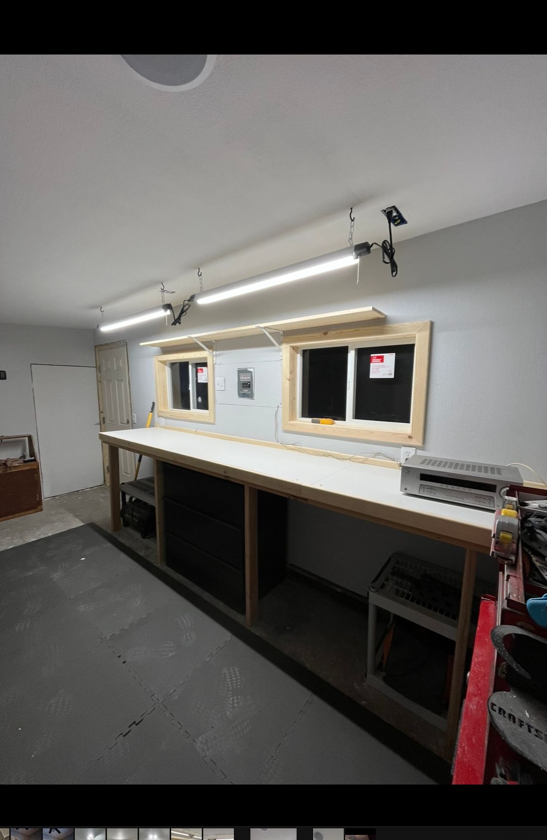 A long, white workbench sits against a gray wall with two small windows and overhead shop lights in a workshop.