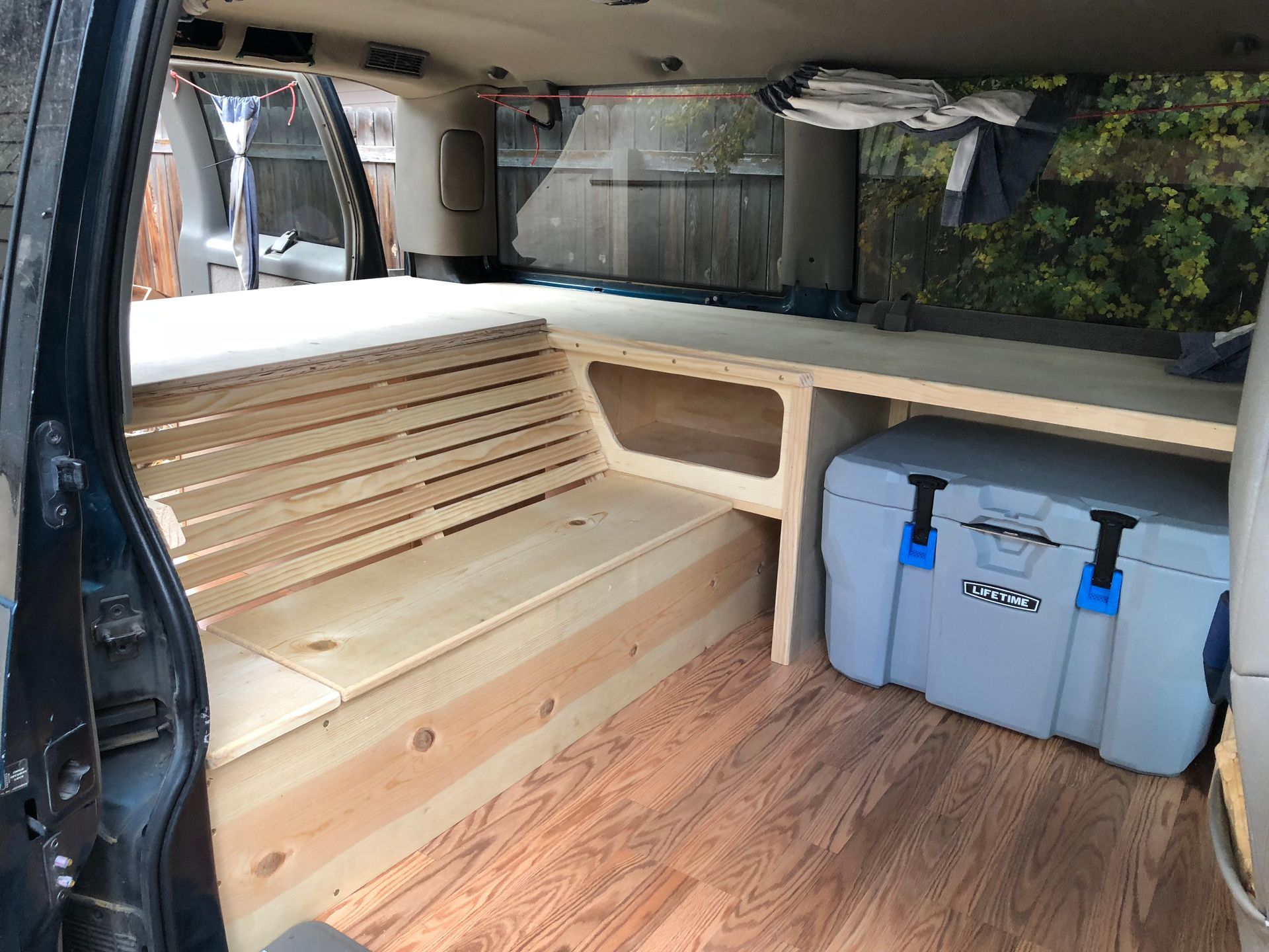 Custom wooden platform bed with slatted base, storage, and a large grey cooler inside a vehicle with wood-look flooring.