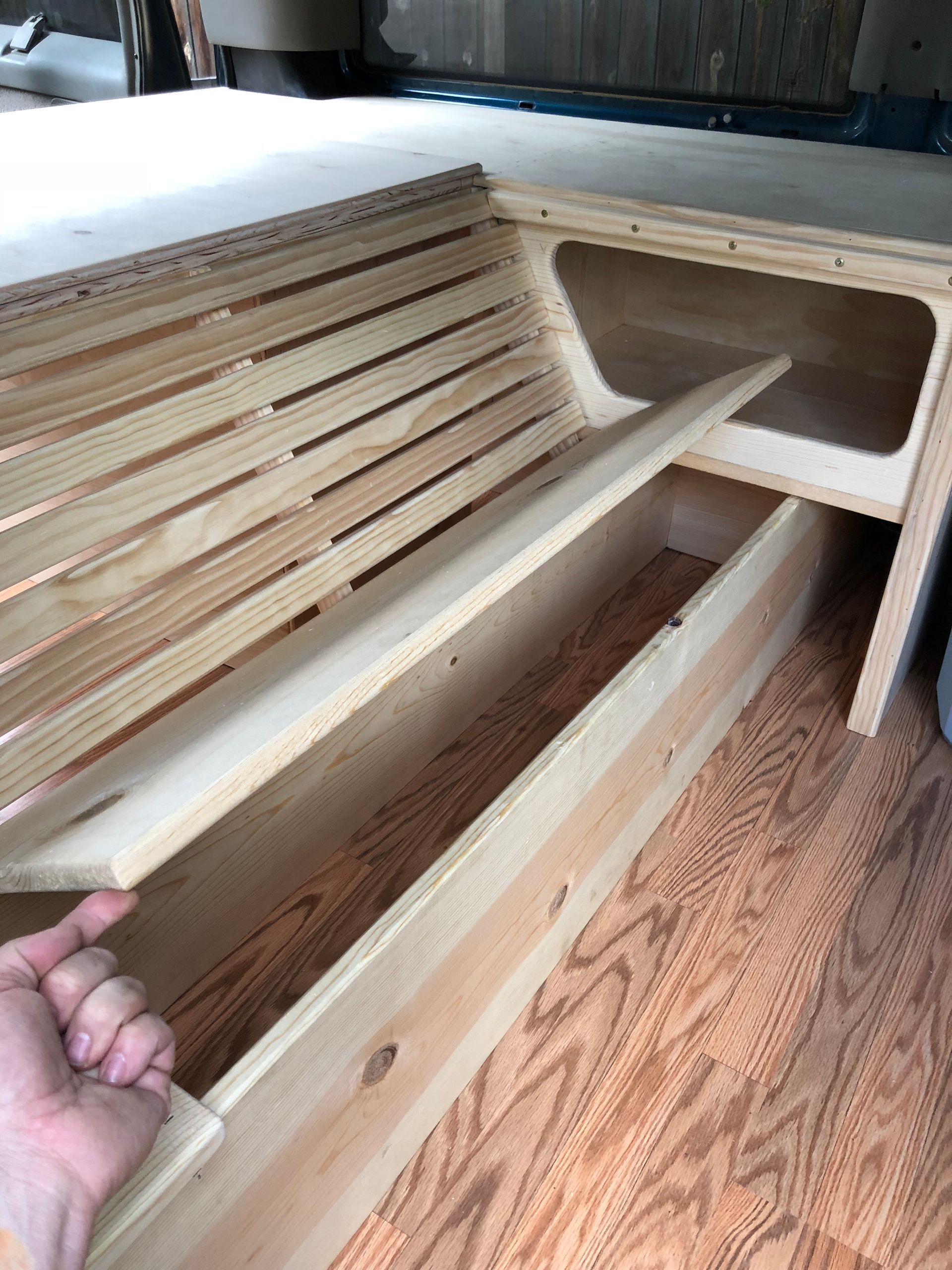 A hand lifts a wooden slat from a custom bed frame being built inside a vehicle with wood-grain flooring.
