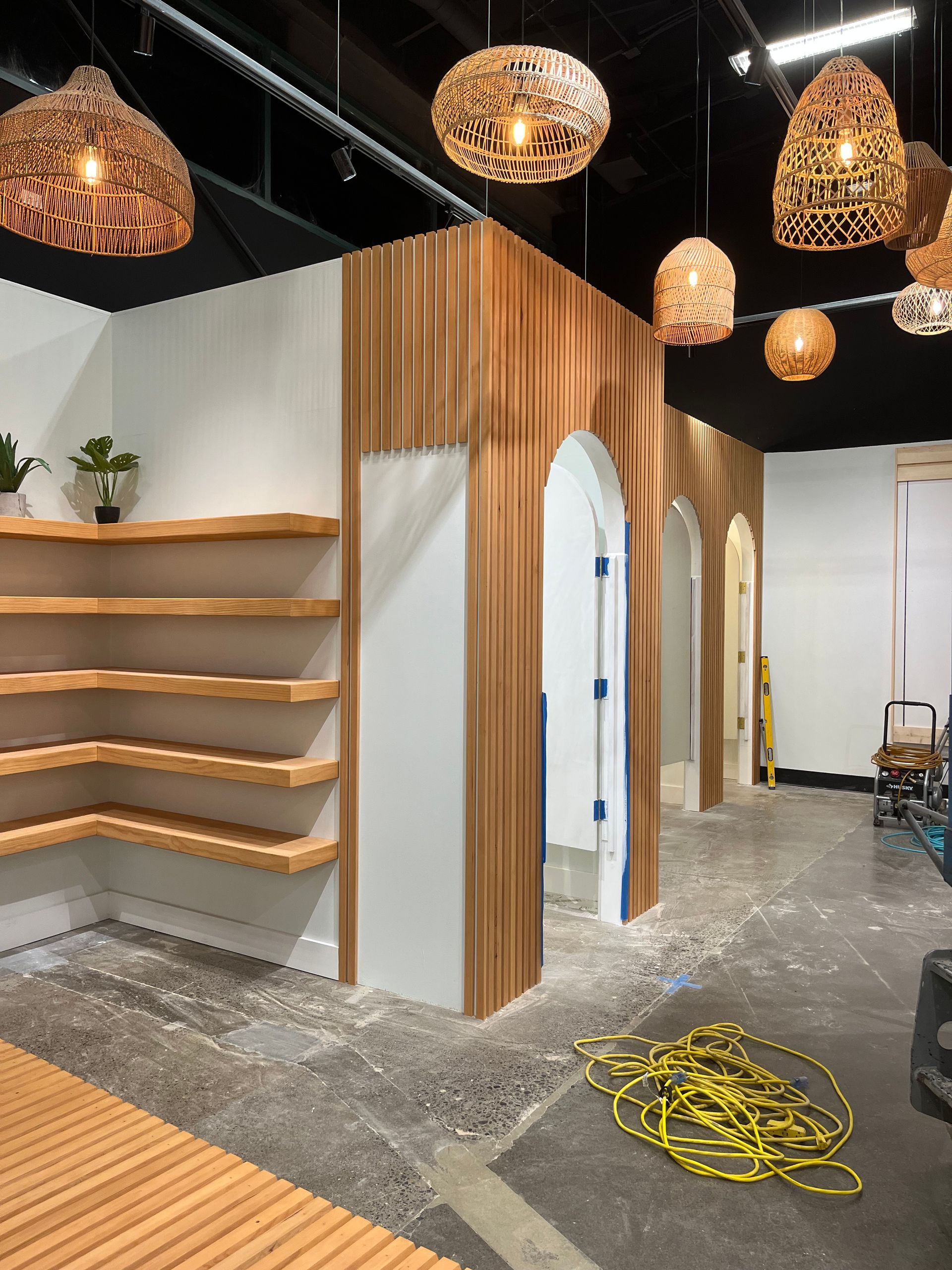 Retail interior under construction with arched wood panel doorways, wall-mounted floating shelves, and pendant lights.