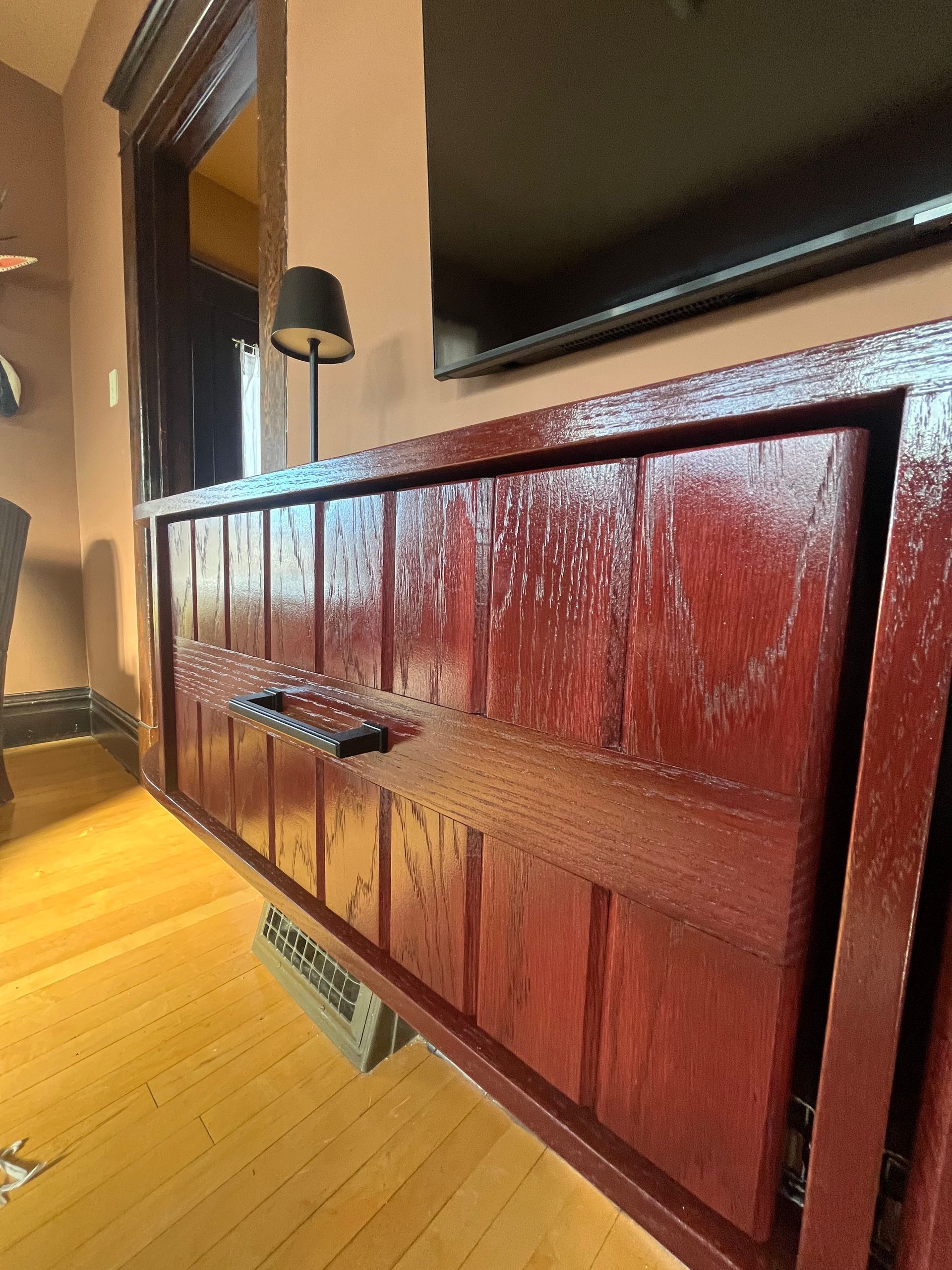 A glossy, reddish-brown wooden cabinet with vertical panels and a dark metal handle, positioned below a wall-mounted TV.