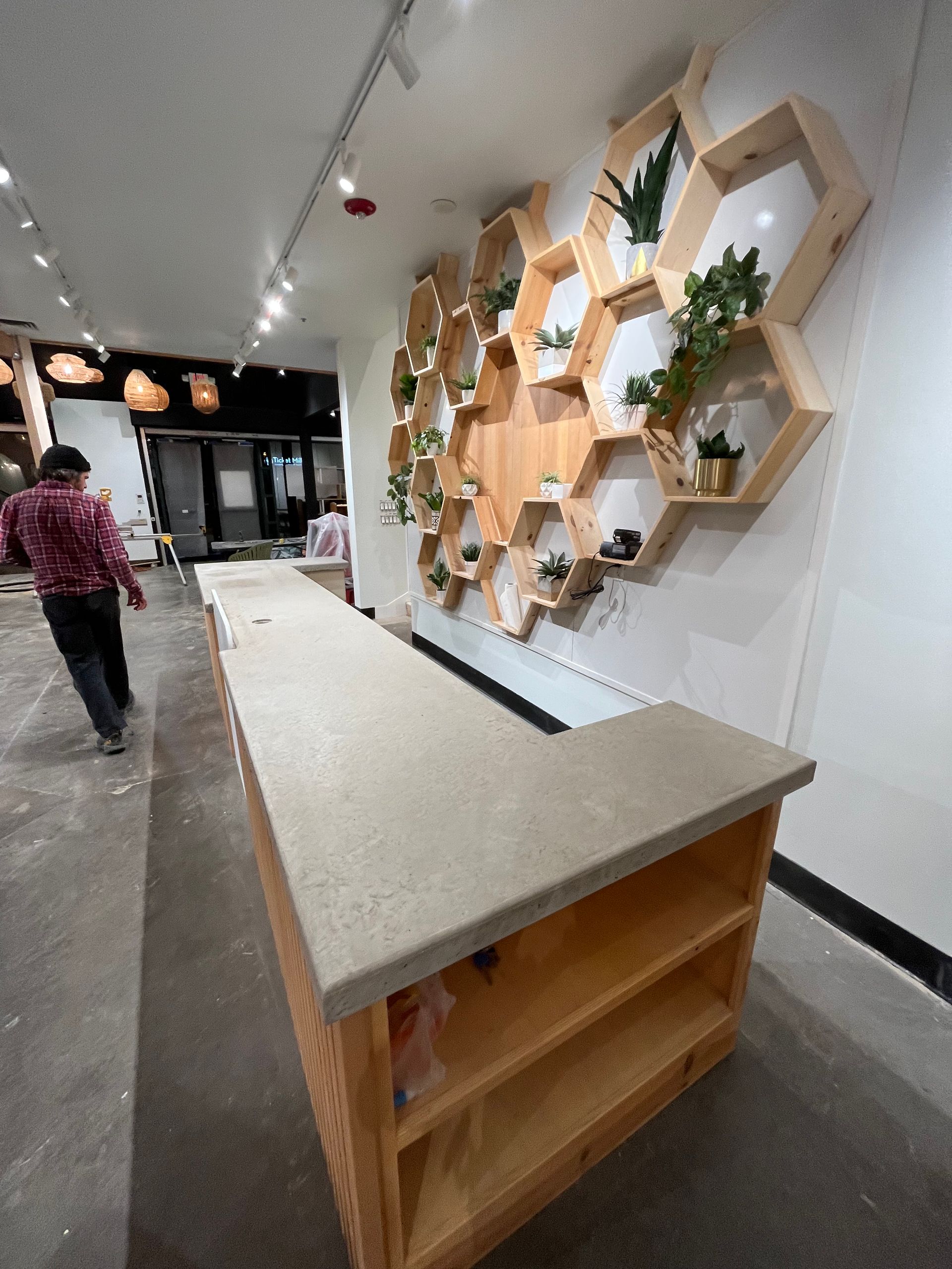 A person walks past a wooden counter next to a large honeycomb-shaped wall display decorated with small potted plants.