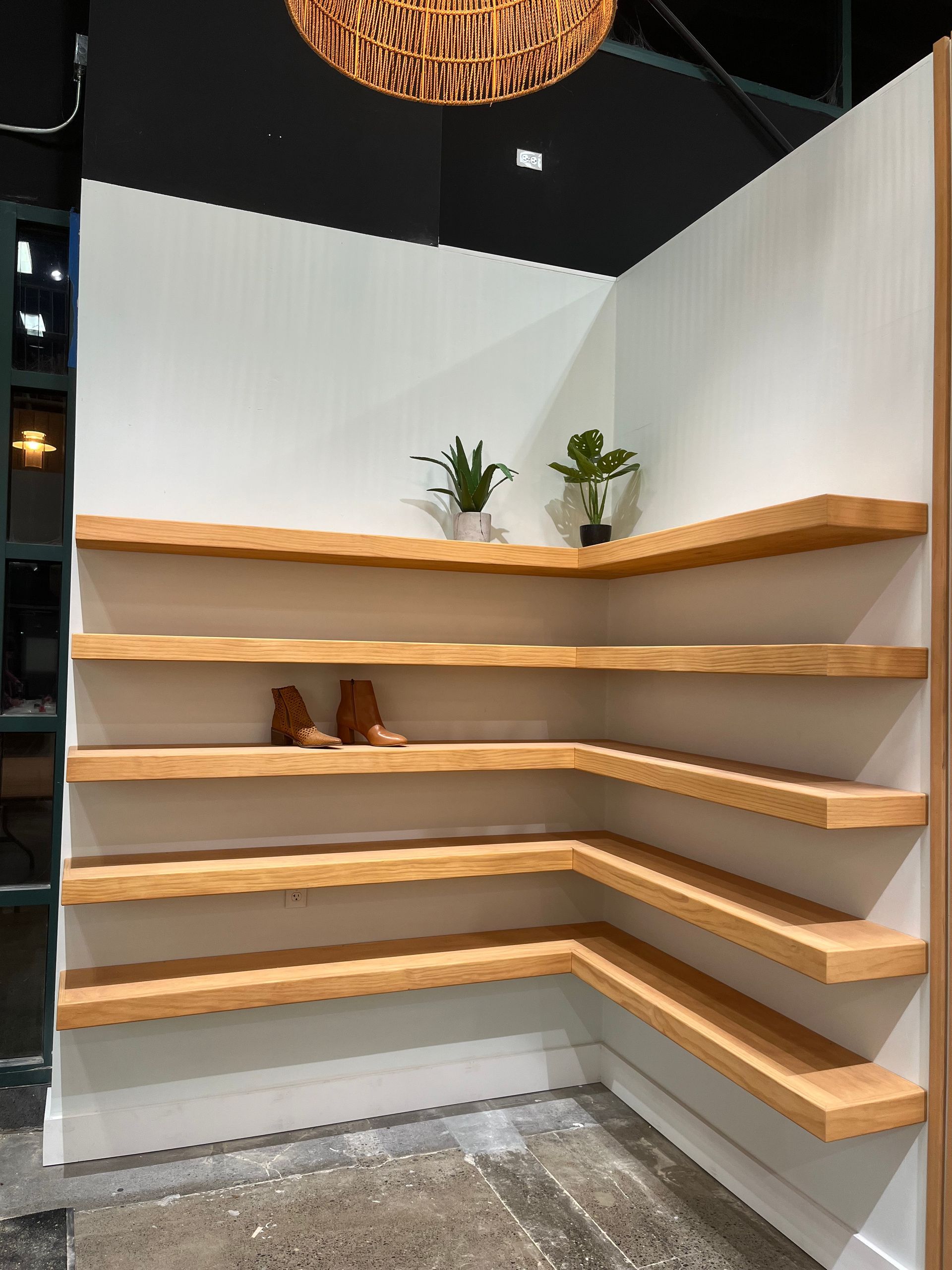 A corner display with five light wood floating shelves mounted on white walls, holding two potted plants and a pair of boots.