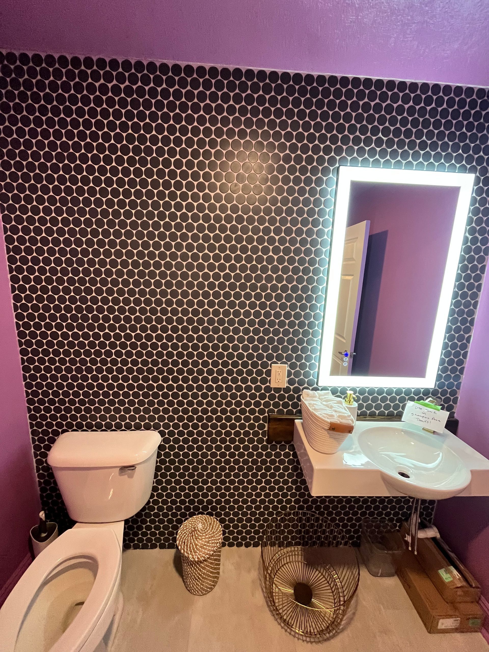 A bathroom with a purple wall, a black-and-white hexagonal tiled accent wall, a white toilet, and a lit vanity mirror.