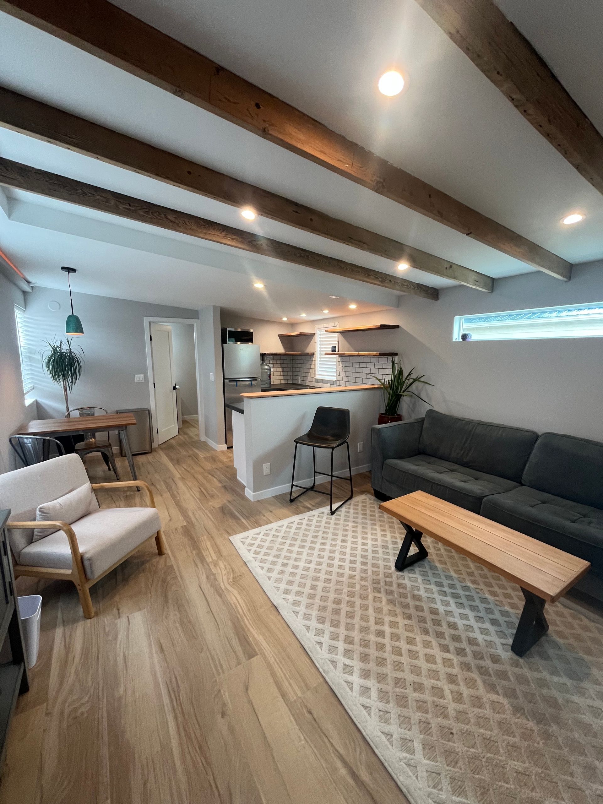 Open-concept living room with wood-beamed ceiling, grey sofa, light-colored chair, wooden coffee table, and kitchen area.