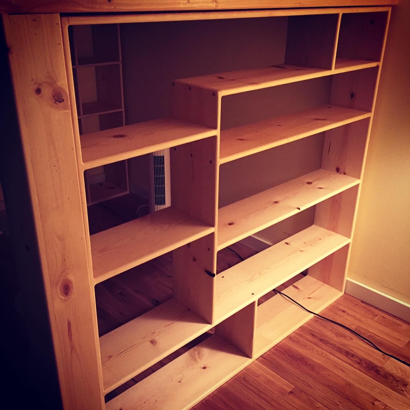 A tall, open-concept wooden bookshelf with a staggered, asymmetric shelf design sitting on a hardwood floor.