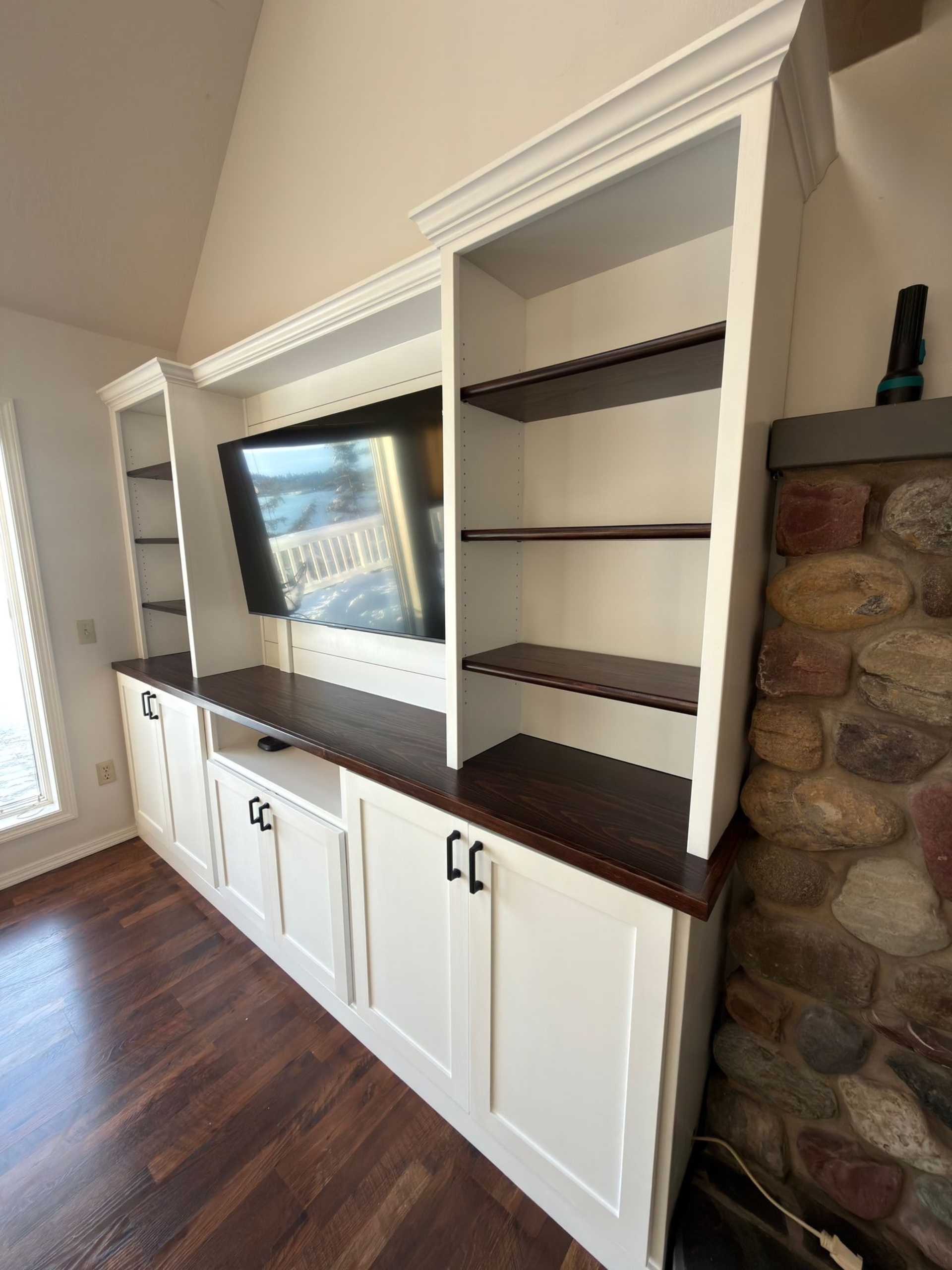 Built-in white cabinets with dark wood shelving and countertops flank a wall-mounted television next to a stone fireplace.