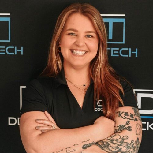 Woman with crossed arms smiles; she has tattoos and stands in front of a company logo.