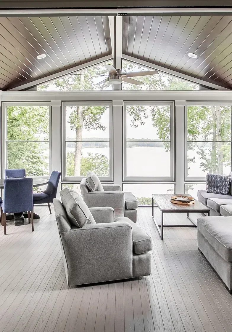 Bright, airy living room with large windows overlooking a lake. Gray furniture, wood ceiling, and light-colored floors.