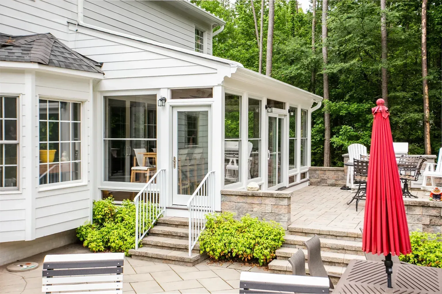 Screened porch with white trim, door, and stairs, leading to a stone patio with a red umbrella.