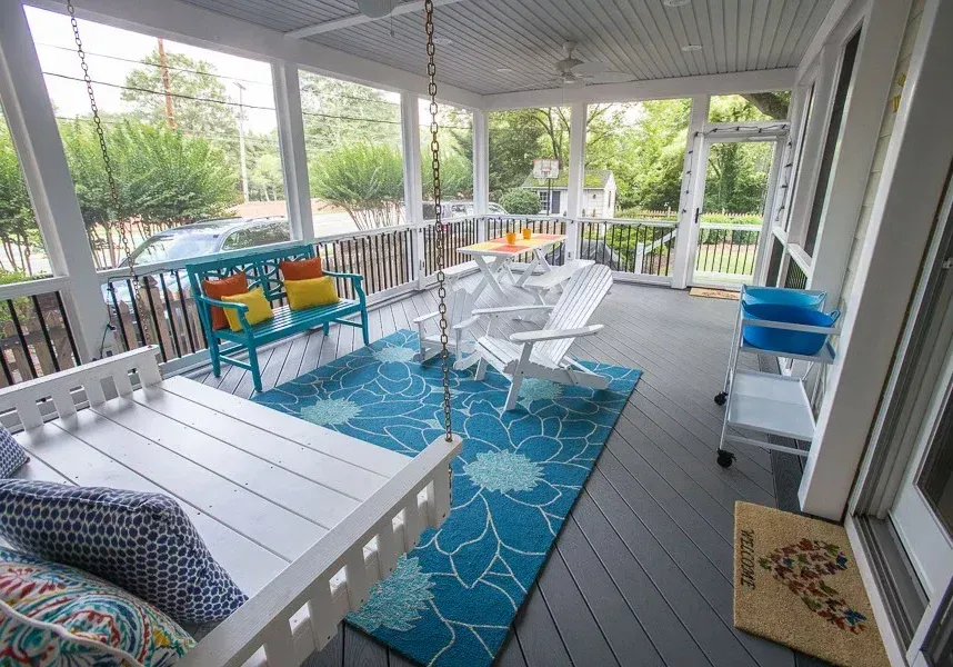 Screened-in porch with blue rug, white swing and Adirondack chairs, teal bench with colorful pillows, and small rolling cart.