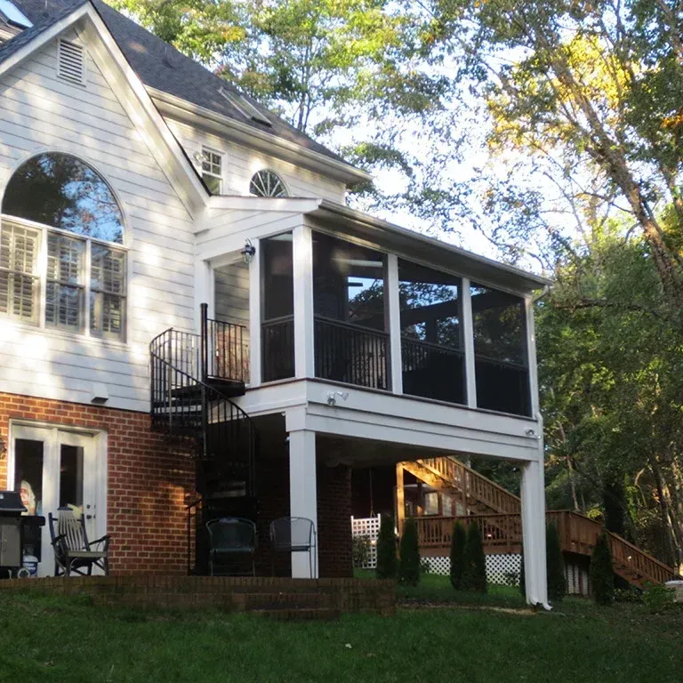 White house with screened porch, spiral staircase, and brick facade, set in a wooded area.