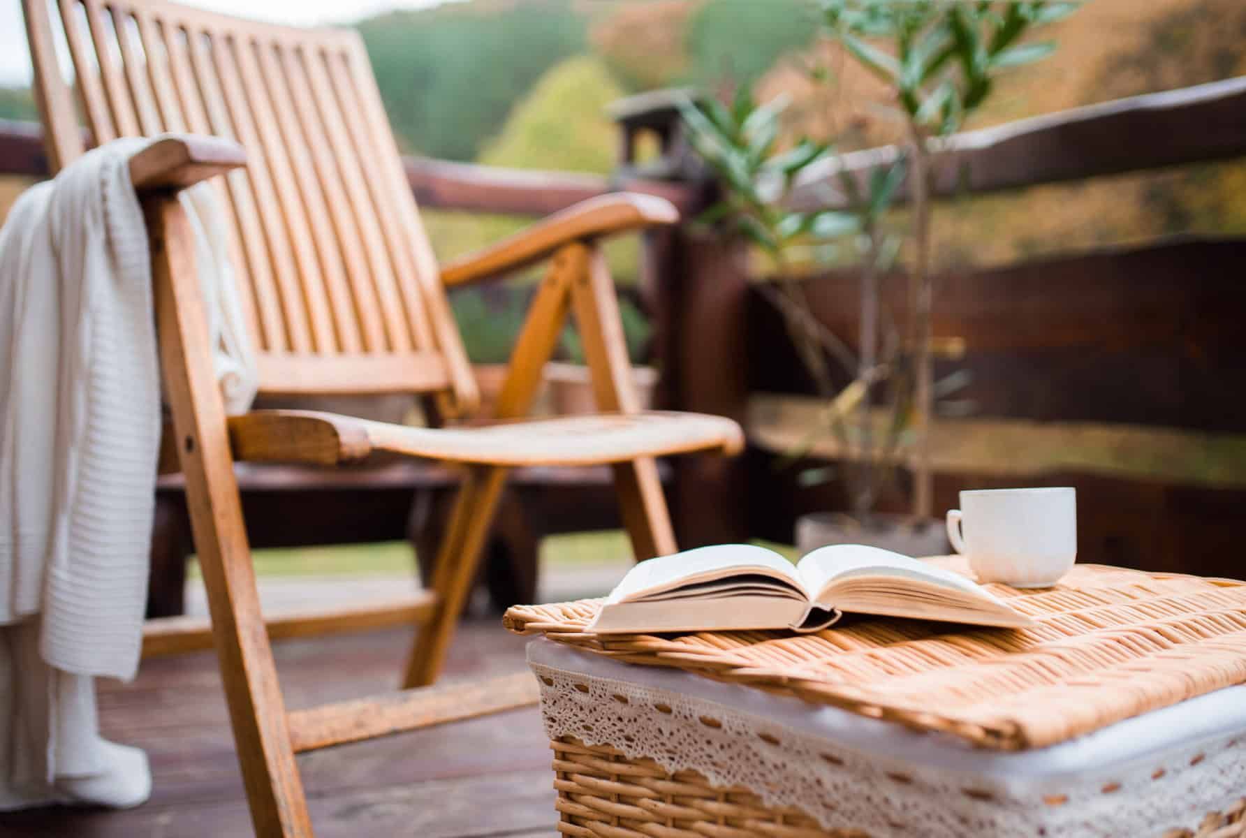 Wooden deck with chair, open book, and coffee cup on wicker table. A white sweater hangs on the chair.