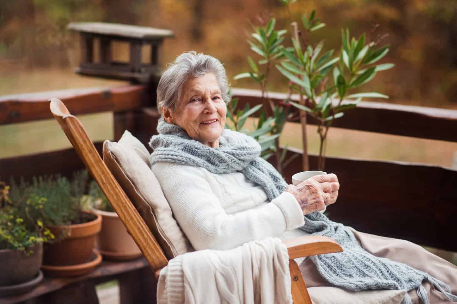 Woman in a rocking chair on a porch, holding a mug. She wears a scarf and smiles. Outdoors with plants.