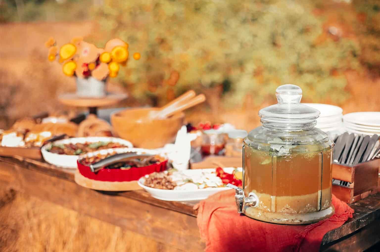 Outdoor buffet with drinks dispenser, food platters, and wooden table. Autumnal colors.