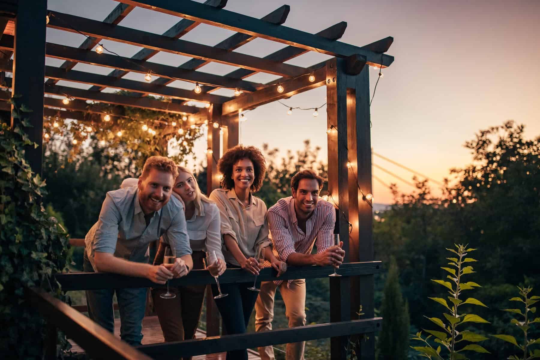 Four friends on a wooden deck with string lights; enjoying sunset with drinks.