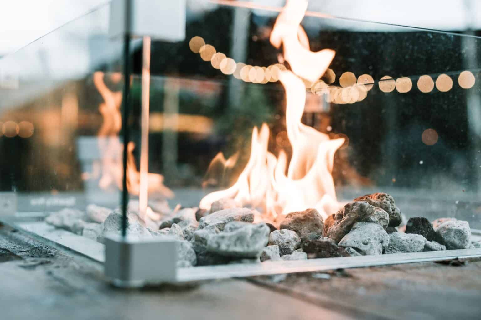 Fire pit with orange flames, gray rocks, and glass enclosure; outdoor setting.
