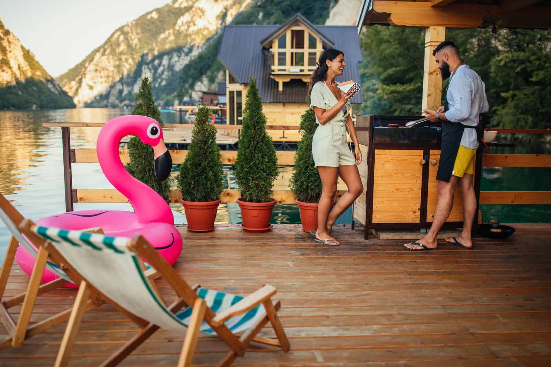 Couple grilling and relaxing on a wooden deck by a lake, mountains in the background, inflatable flamingo.