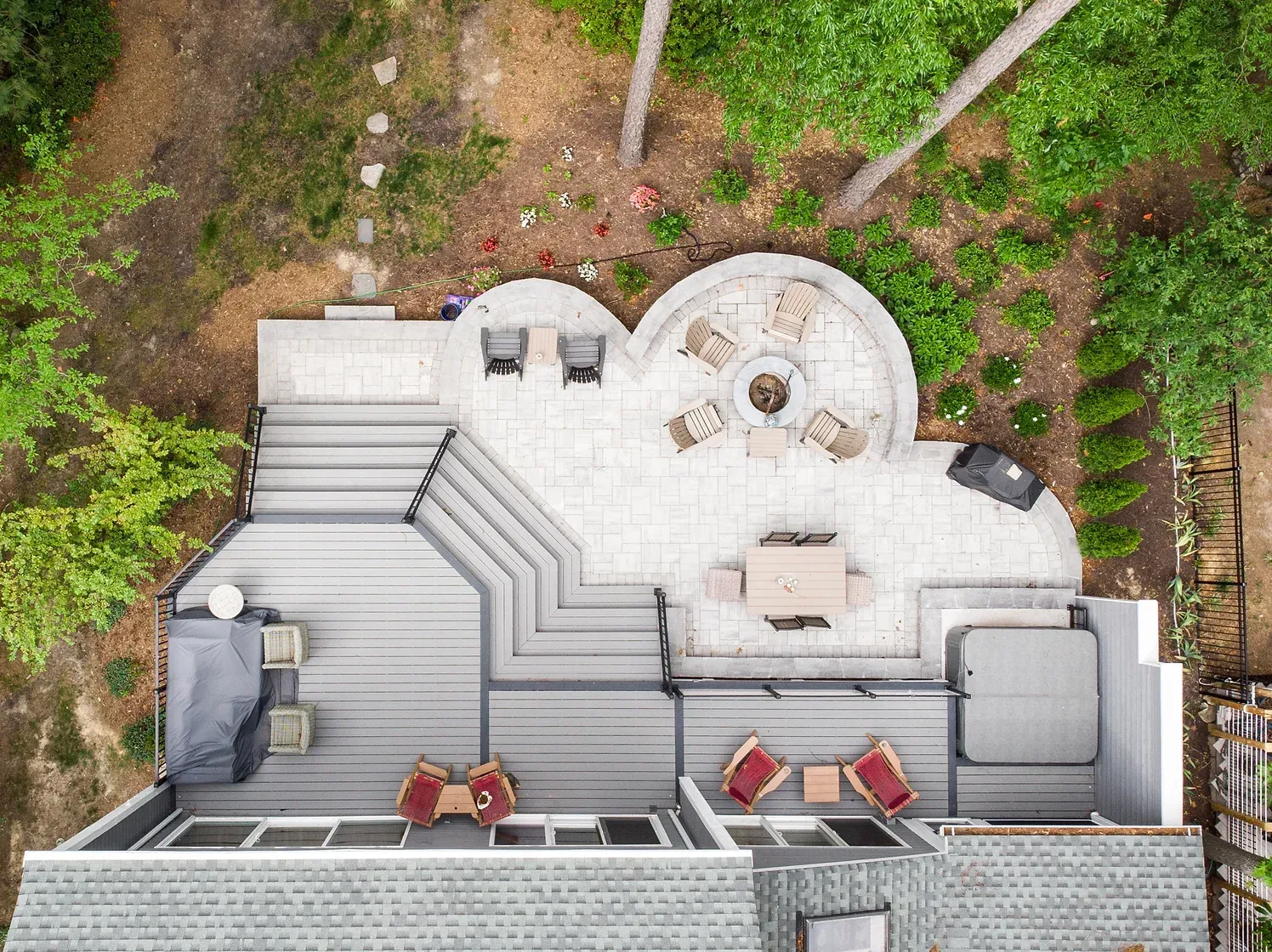 Overhead view of a home with a gray deck, patio with fire pit, and hot tub surrounded by trees.