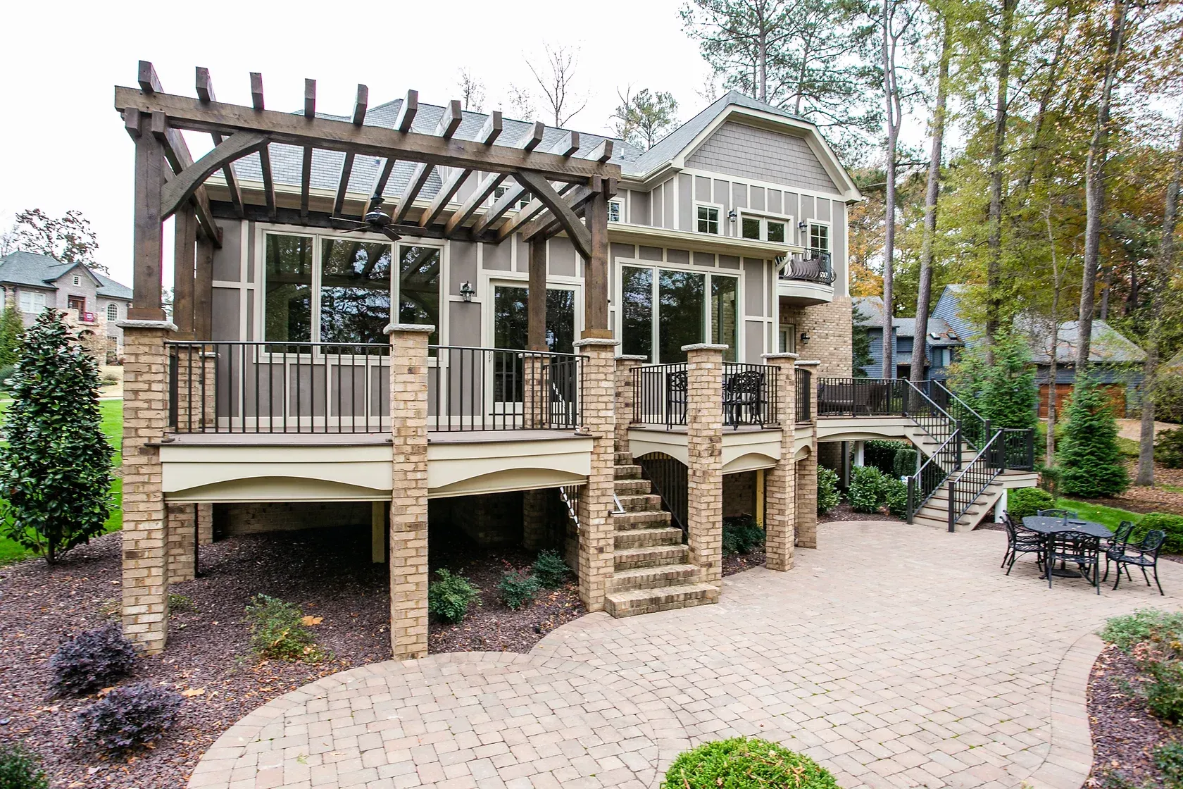 Backyard deck with pergola, brick columns, stairs, and stone patio. Brown siding, green trees, overcast sky.