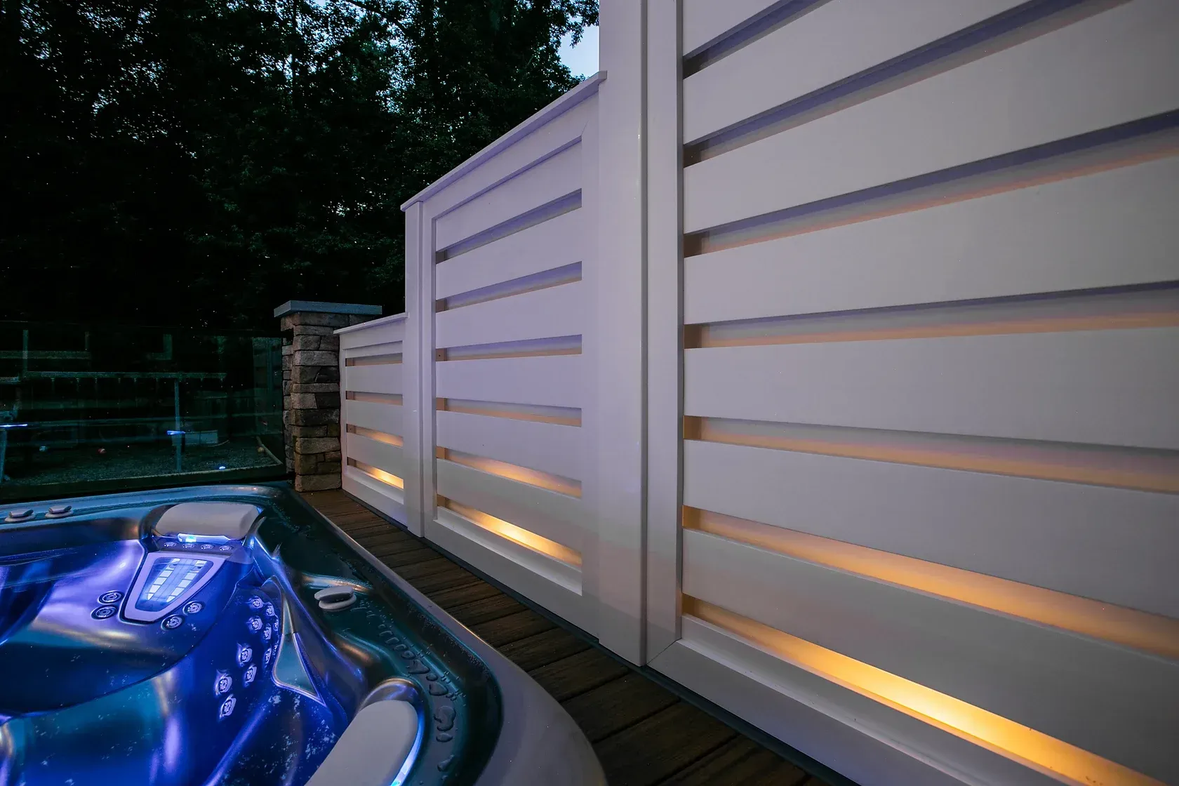 Hot tub lit with blue lights next to a white slatted fence with warm accent lighting.