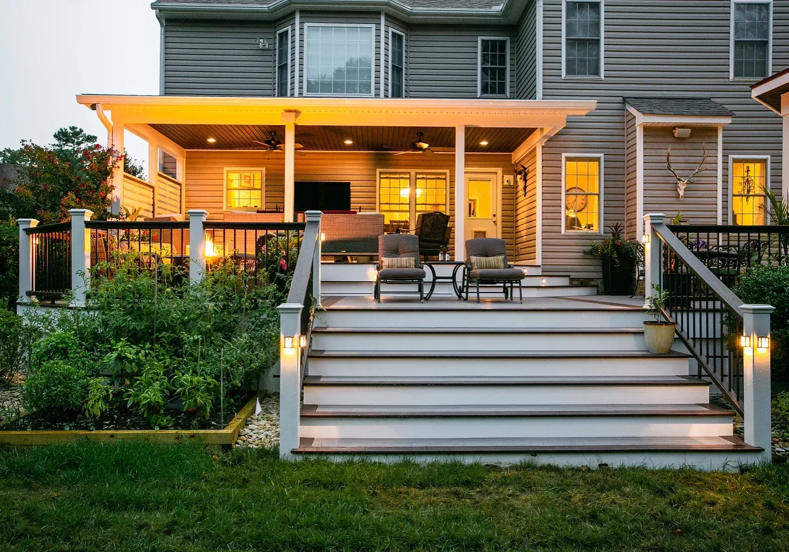 Backyard deck with built-in stairs, seating, and overhead lighting. Gray siding and lush greenery surround.