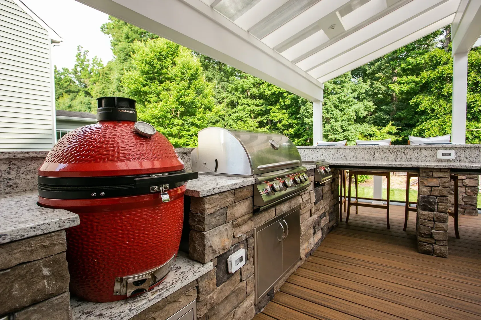 Outdoor kitchen with a red ceramic grill, stainless steel grill, and stone countertop under a white pergola.