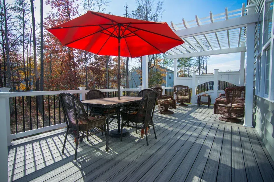 Patio with table, chairs, red umbrella, and pergola. Deck overlooks trees and blue sky.