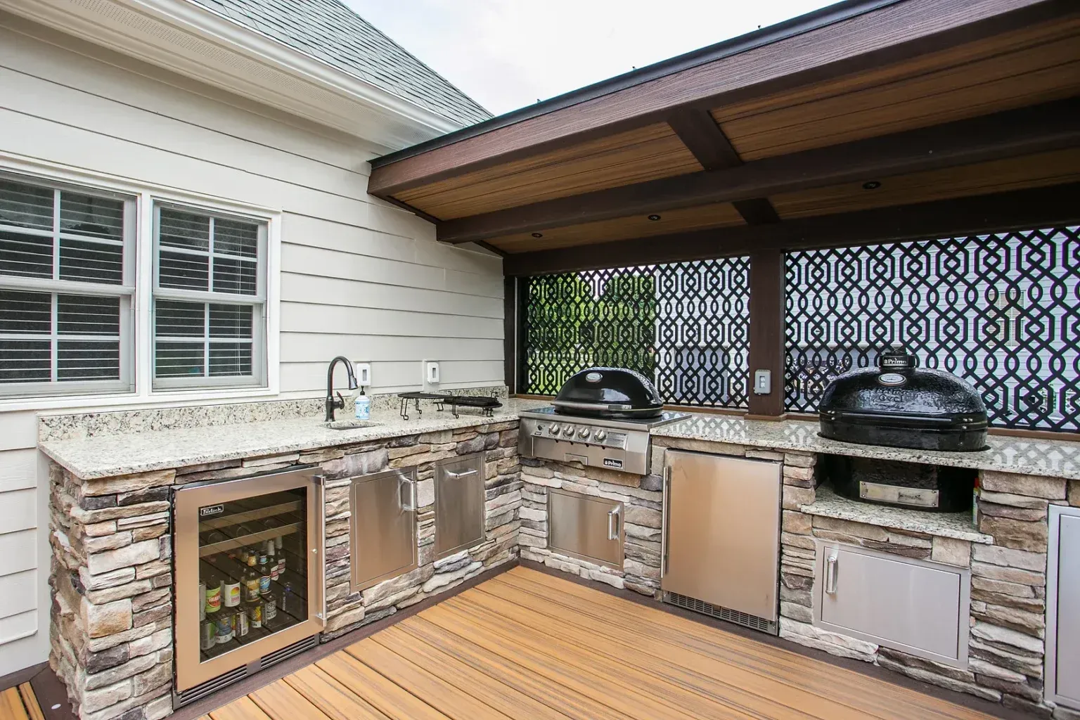 Outdoor kitchen with stone and stainless steel, featuring grill, fridge, and sink under a wooden canopy.