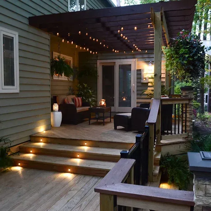 Wooden deck with steps lit by lights, leading to a seating area under a pergola strung with lights.