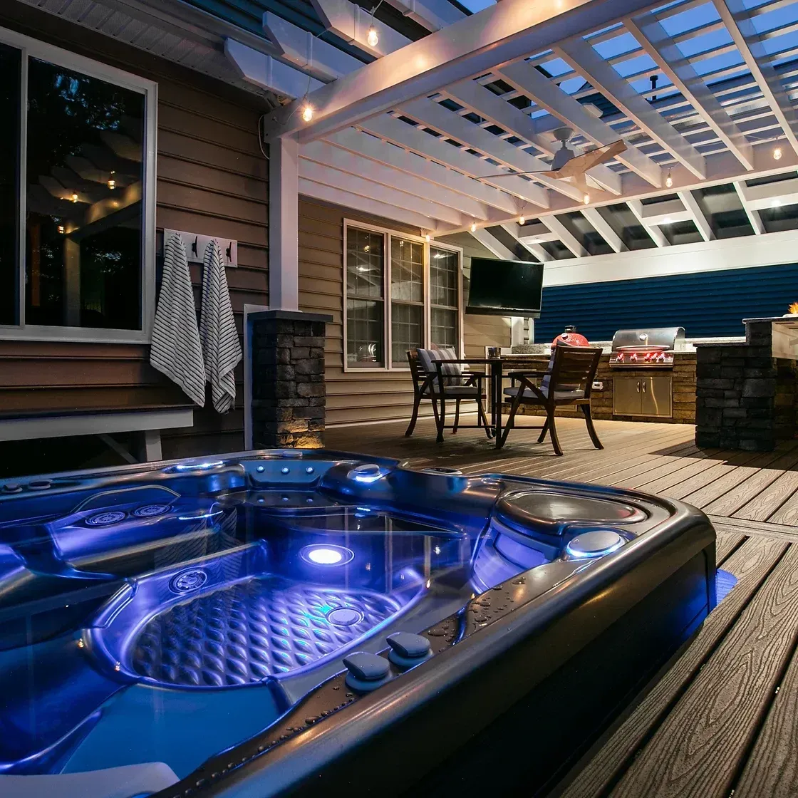Outdoor deck with lit hot tub, dining set, grill, and pergola at dusk.