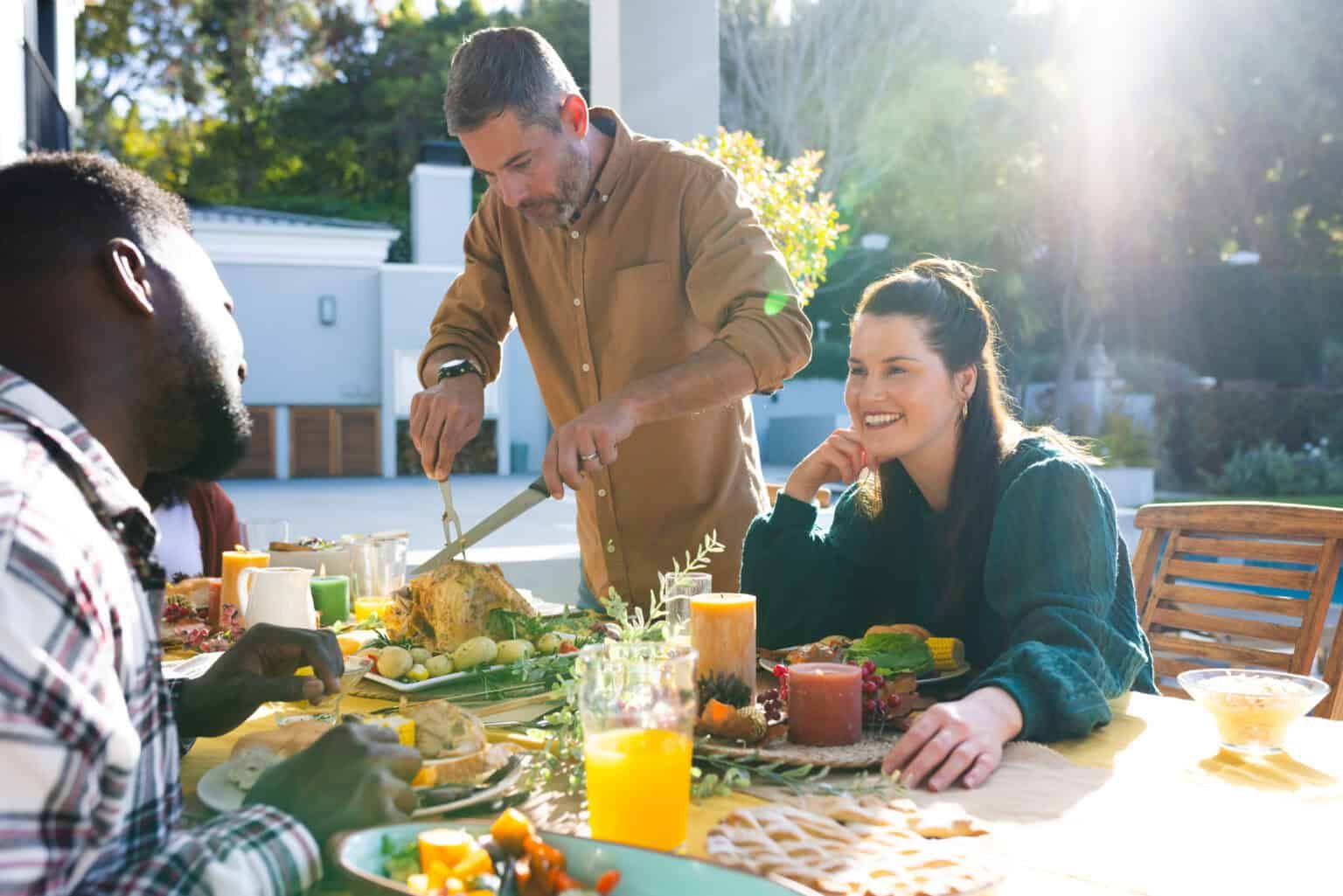 People at outdoor table, man carving turkey, woman smiling, sunlight.