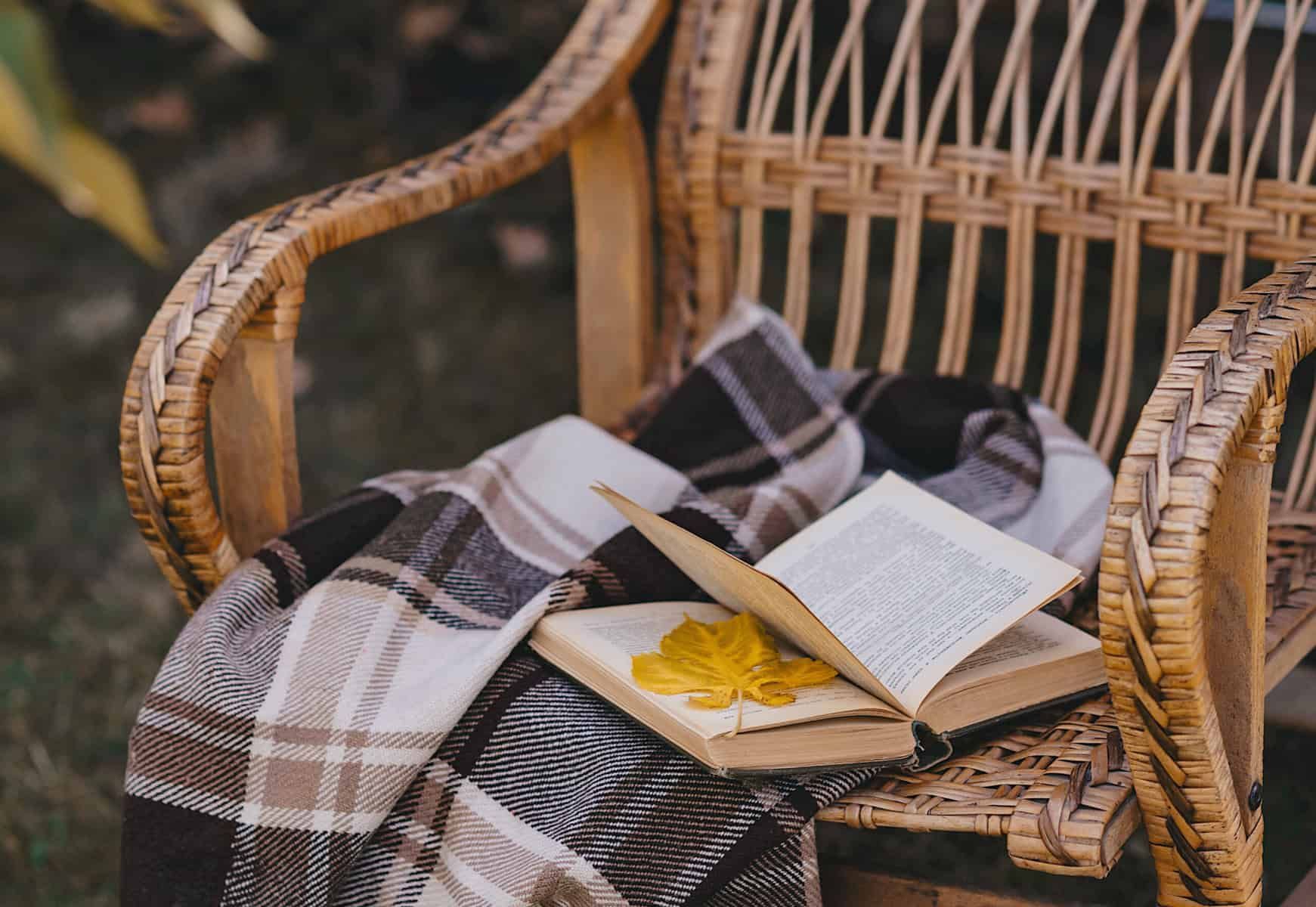 Wicker chair with plaid blanket, open book with a leaf, outdoors.
