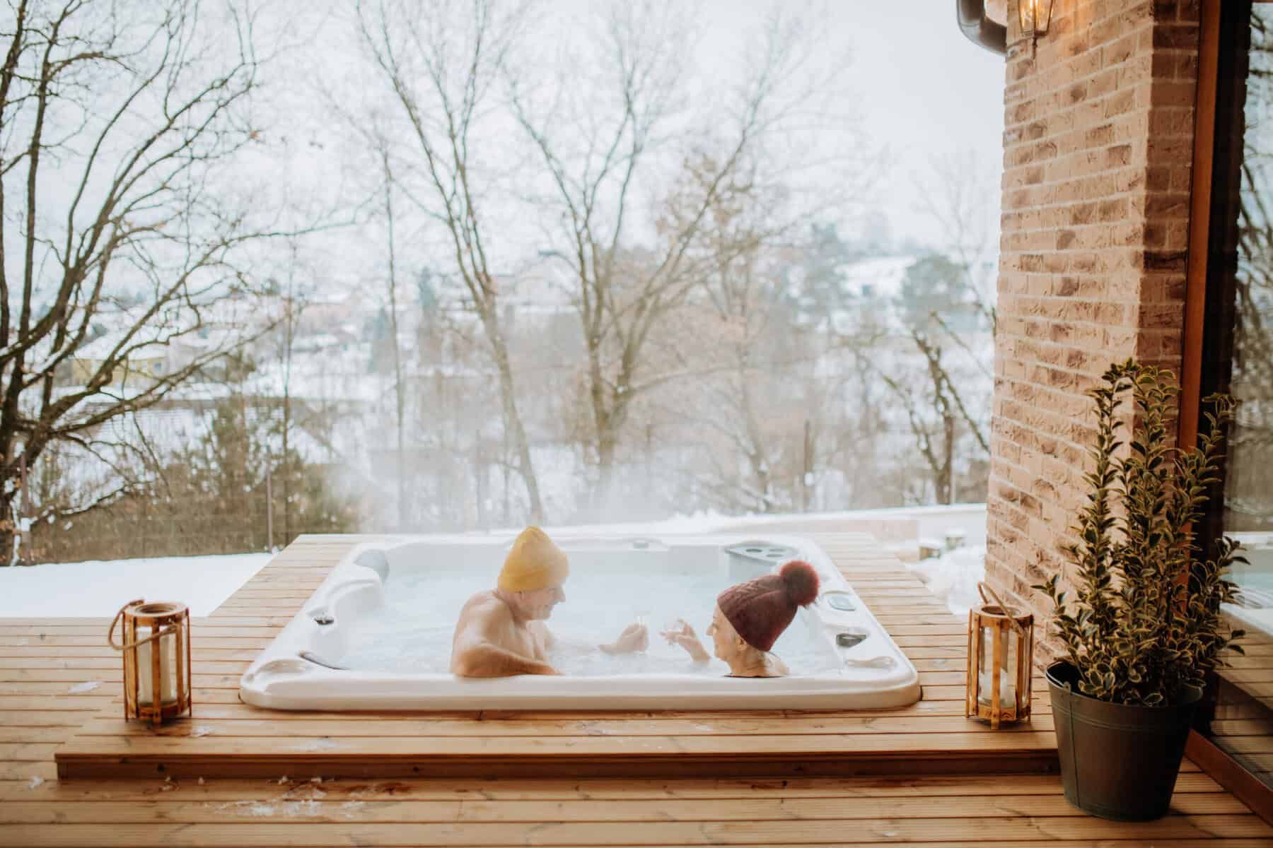 Two people wearing hats sit in a steaming hot tub on a snowy deck, overlooking a winter landscape.