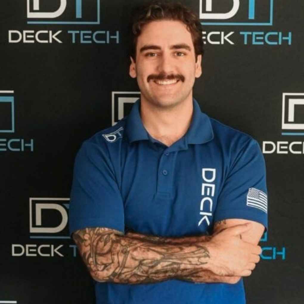 Man with mustache in blue polo shirt with arms crossed, standing in front of a backdrop with