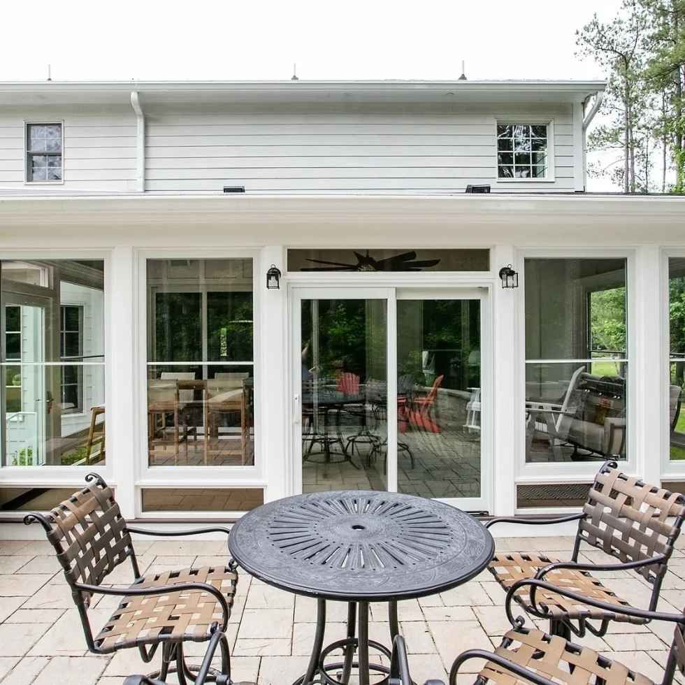 Patio with table, chairs, and sliding glass doors leading into a sunroom. White house in background.