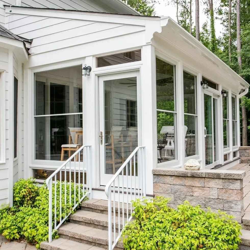White sunroom with glass windows and door, stone patio, white railing and steps. Green bushes surround.