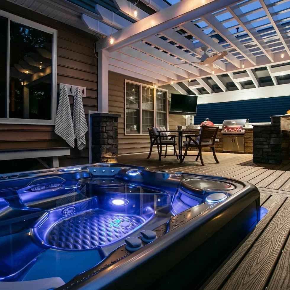 Outdoor deck with illuminated hot tub, pergola, grill, and seating. Blue, brown, and white color scheme.