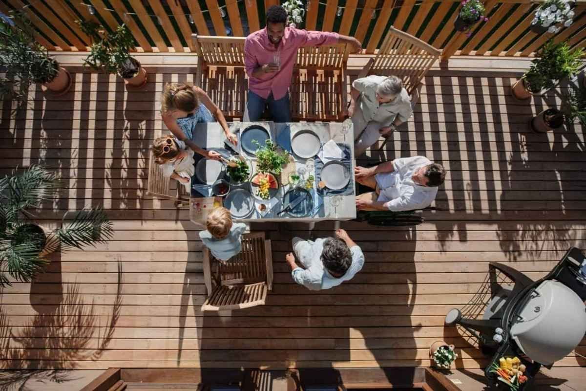 Family gathering around a table on a wooden deck, preparing a meal. Sunlight, greenery, and a grill are visible.