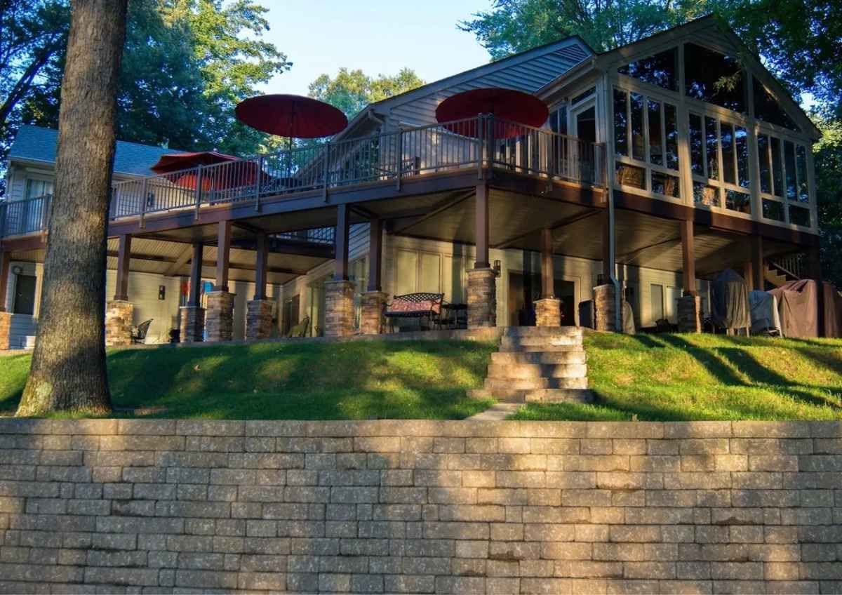 Two-story house with deck and sun umbrellas. Overlooks a yard, retaining wall, and trees.