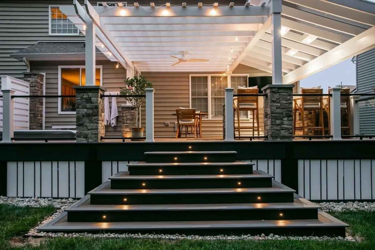 Deck with stairs illuminated by lights, under a pergola. Backyard with a house in the background.