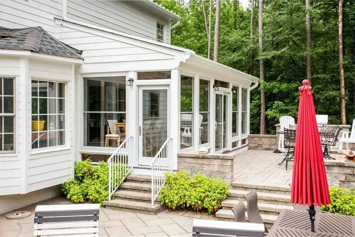 Screened porch with white trim, door, and stairs, leading to a stone patio with a red umbrella.