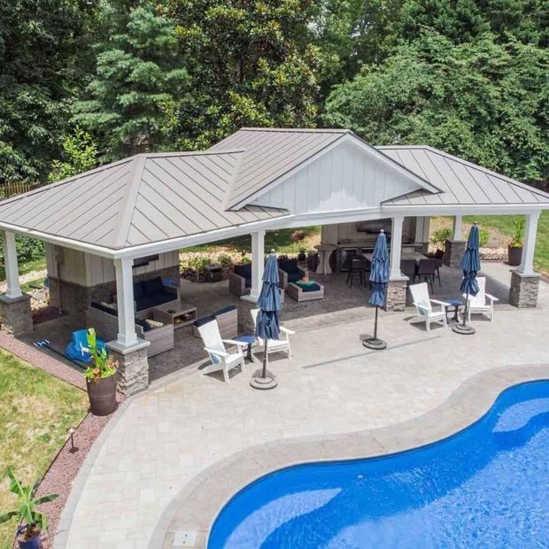 Outdoor patio with pool. White columns, grey roof, seating areas, blue umbrellas, and a curved pool with clear water.